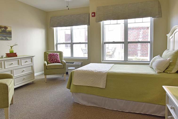 A cozy bedroom with a bed, two chairs, a dresser, and two large windows letting in natural light.