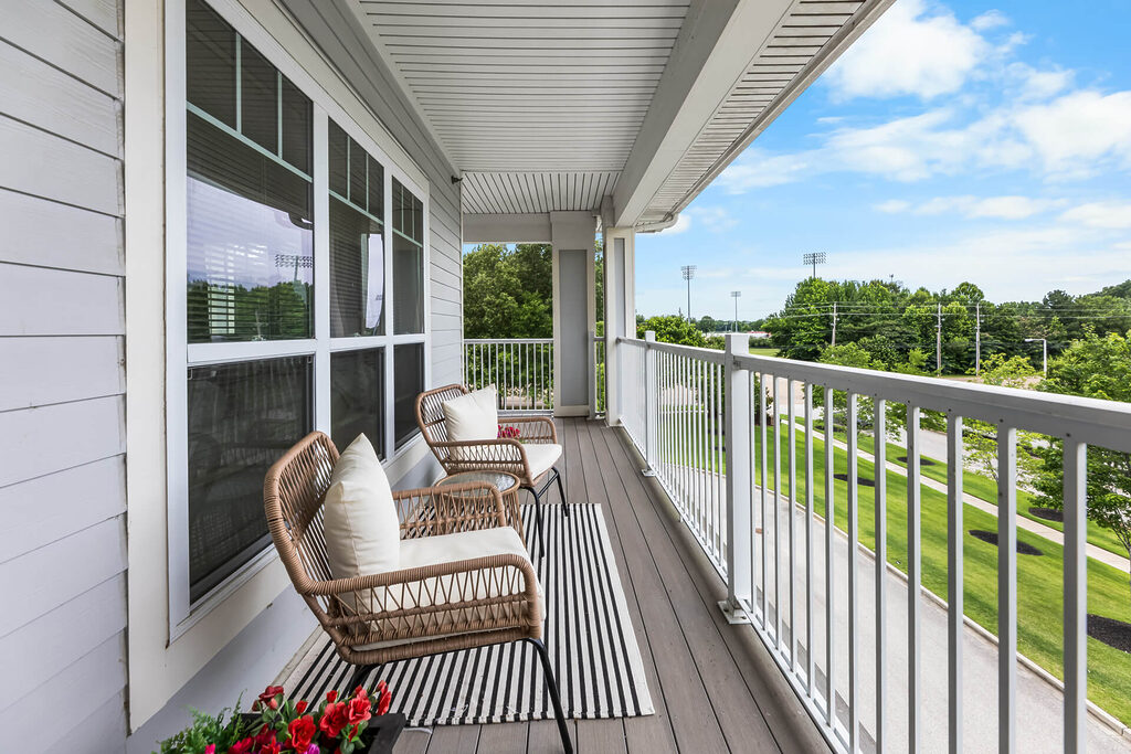 Outdoor balcony with two chairs, striped rug, and potted flowers overlooking green area.